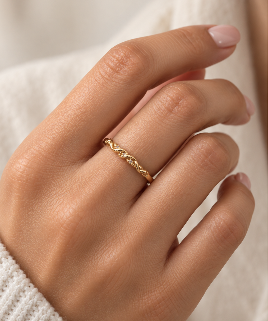 Gold-tone ring with a slender band, flowing interlacing wave pattern and small clear reflective accents, displayed on a white background and worn on a finger.