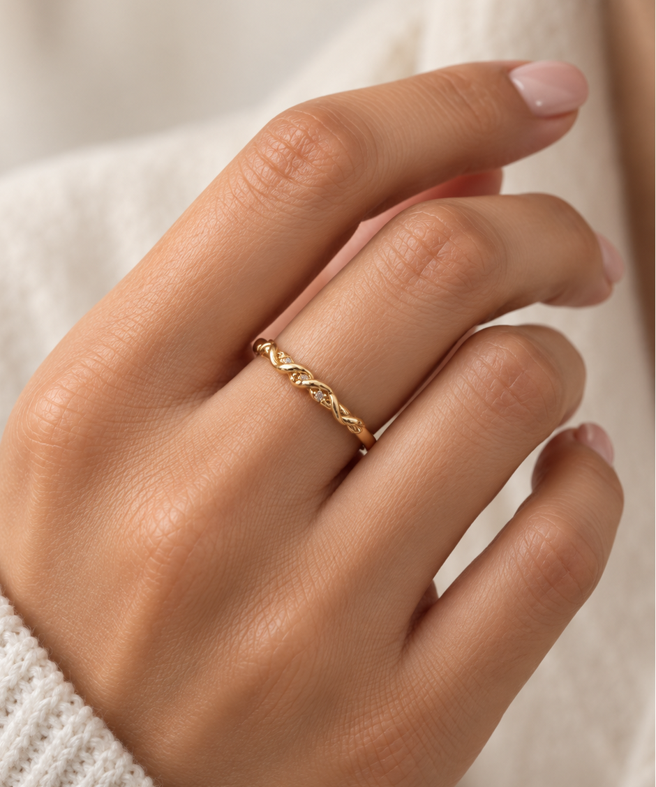 Gold-tone ring with a slender band, flowing interlacing wave pattern and small clear reflective accents, displayed on a white background and worn on a finger.