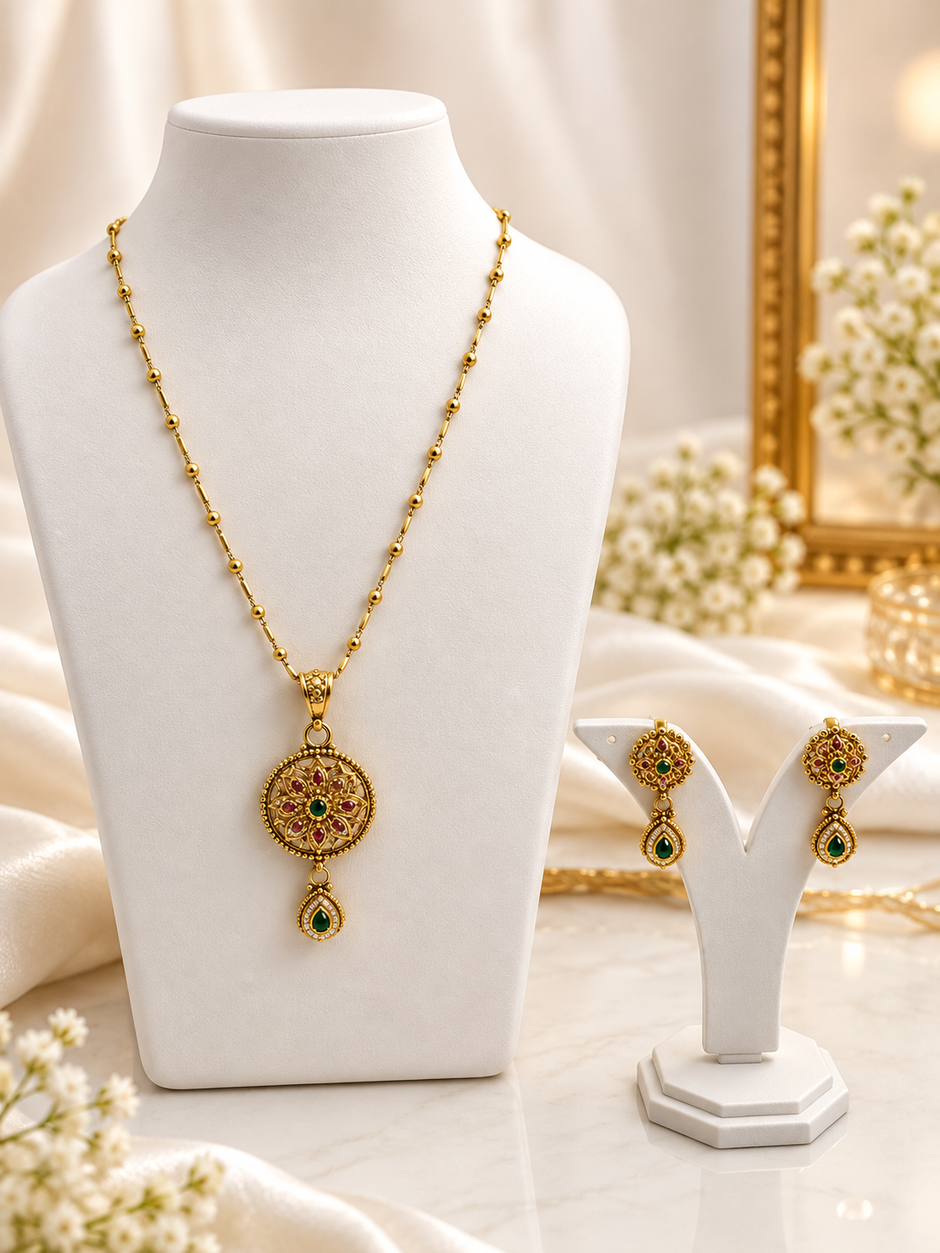 Gold-tone floral pendant necklace set displayed on white jewellery stands, featuring green and magenta-pink accents, a beaded chain, and matching earrings with green teardrop-shaped drops.