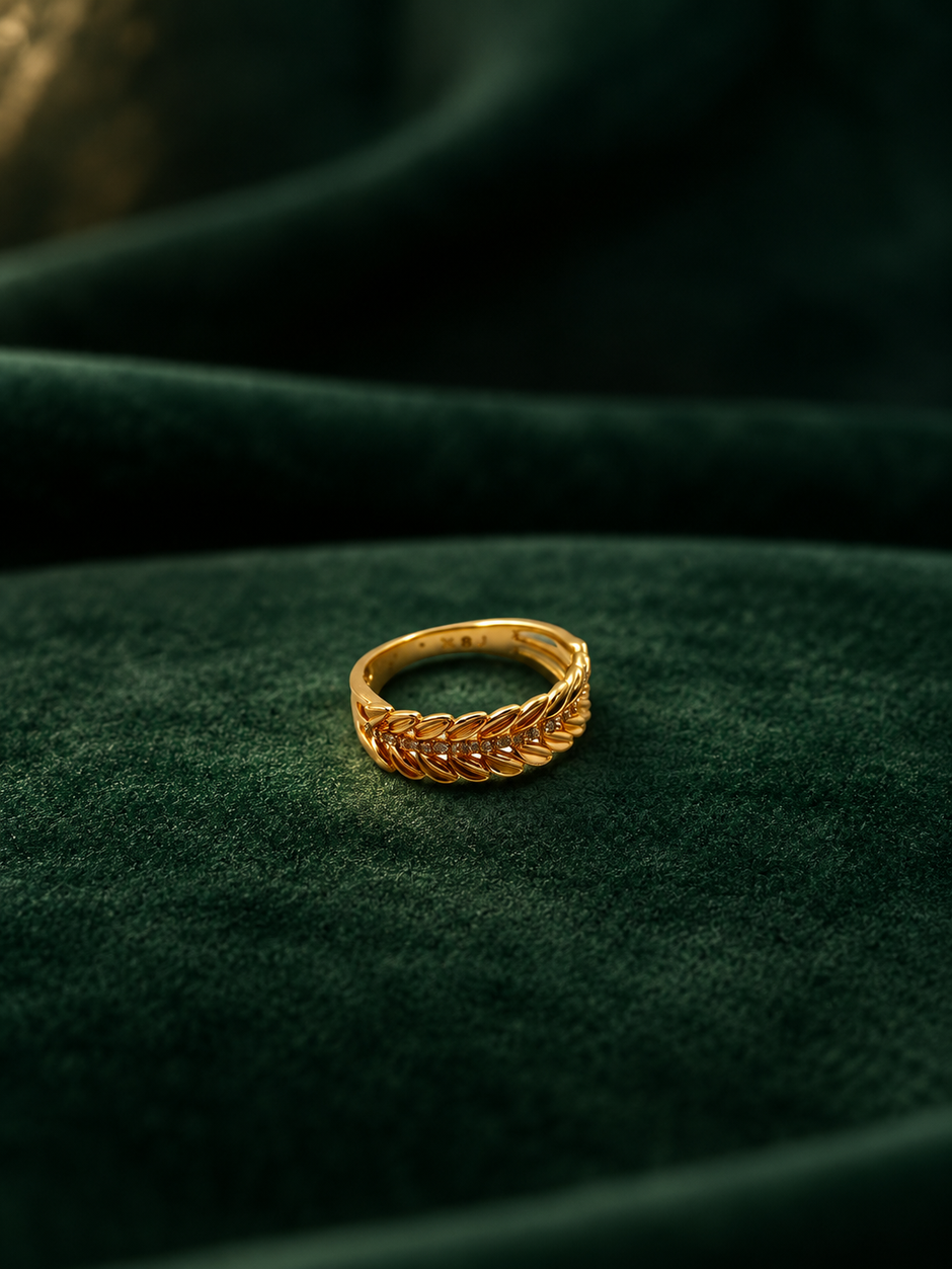 Gold-tone decorative ring with overlapping diagonal leaf-like curves and a central row of small clear stones, shown on a white background and worn on a finger.