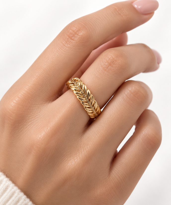 Gold-tone decorative ring with overlapping diagonal leaf-like curves and a central row of small clear stones, shown on a white background and worn on a finger.