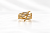 Gold-tone multi-row crossover ring with intertwined curved bands, round clear stones and dark accent detailing on a white background.
