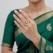 A gold ring with pink floral detailing is worn on a woman's hand, who is also wearing a green saree with gold borders.