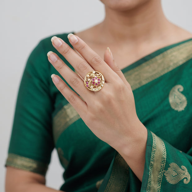 A gold ring with pink floral detailing is worn on a woman's hand, who is also wearing a green saree with gold borders.