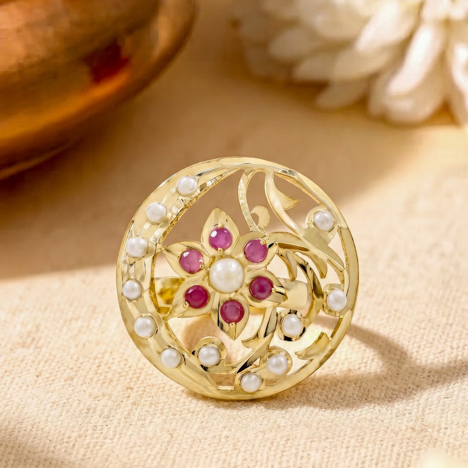A round gold-tone brooch featuring a floral motif with pinkish-red cabochon stones and white faux pearls, displayed on a textured beige fabric background.