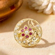 A round gold-tone brooch featuring a floral motif with pinkish-red cabochon stones and white faux pearls, displayed on a textured beige fabric background.