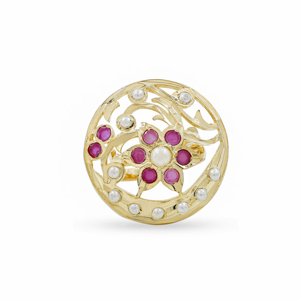 A round gold filigree brooch featuring a central floral motif with pinkish-red gemstones and white pearl-like accents, set against a plain white background.