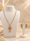 Gold-tone floral halo necklace set displayed on white jewellery stands, featuring a round pendant with green and magenta-pink accents, a decorative beaded chain, and matching earrings with green teardrop-shaped drops.