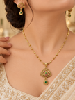 Gold-tone floral temple necklace set displayed on white jewellery stands, featuring a triangular ornamental pendant with green and magenta-pink accents, a beaded chain, and matching earrings with green teardrop-shaped drops.