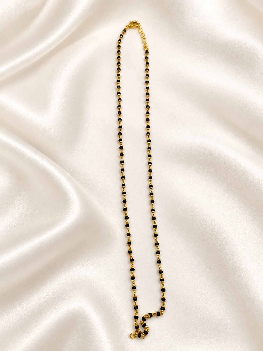 Black beaded necklace with small gold-tone connecting elements and a gold-tone clasp, displayed as an elongated chain on a white background and held in a white-gloved hand. - bhavmayi