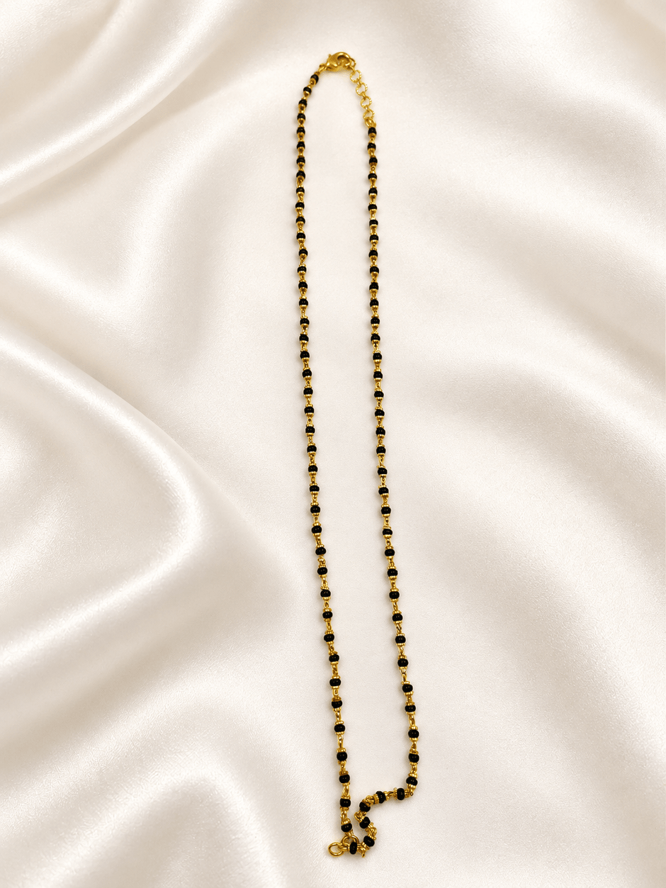 Black beaded necklace with small gold-tone connecting elements and a gold-tone clasp, displayed as an elongated chain on a white background and held in a white-gloved hand. - bhavmayi