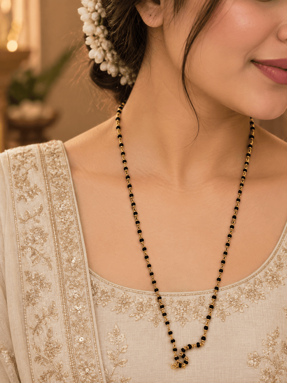 Black beaded necklace with small gold-tone connecting elements and a gold-tone clasp, displayed as an elongated chain on a white background and held in a white-gloved hand. - bhavmayi