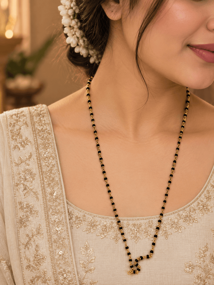 Black beaded necklace with small gold-tone connecting elements and a gold-tone clasp, displayed as an elongated chain on a white background and held in a white-gloved hand. - bhavmayi