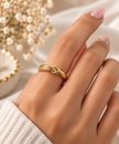 Gold-tone knot ring featuring intertwined curved loops, a row of small clear stones and dark accents, shown on a finger and from the side.