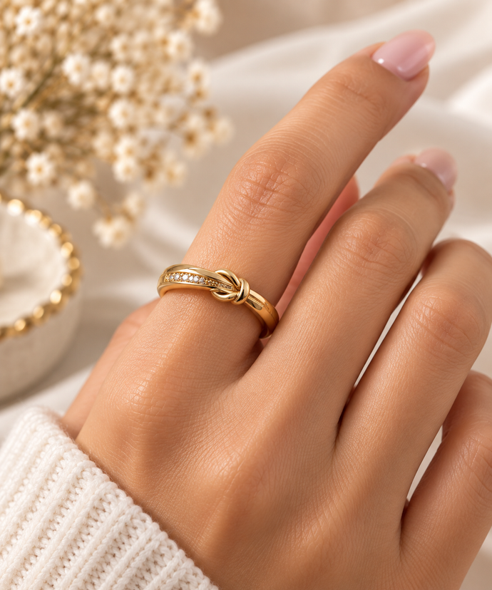 Gold-tone knot ring featuring intertwined curved loops, a row of small clear stones and dark accents, shown on a finger and from the side.