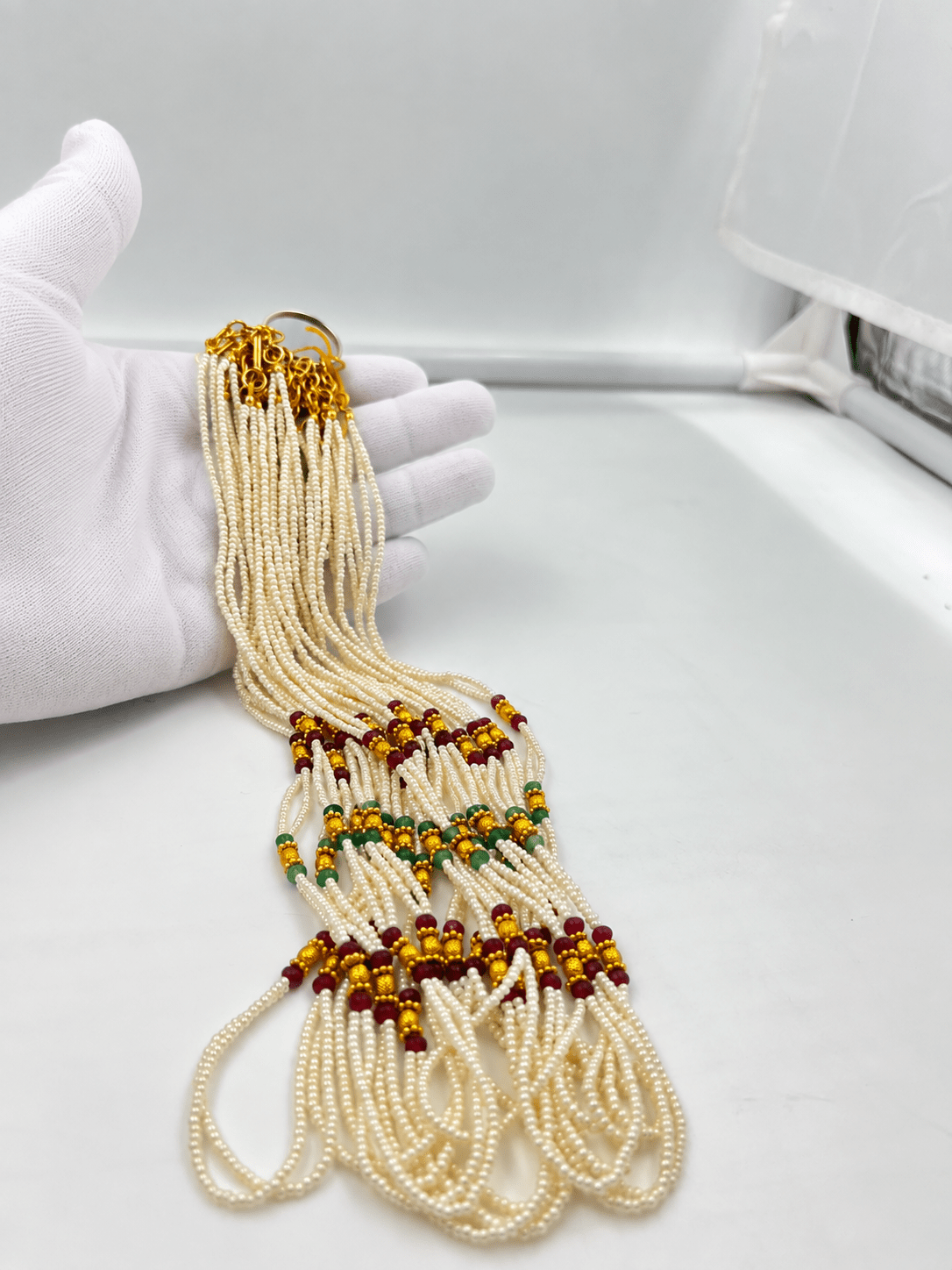 Double-strand white beaded necklace with deep red and green bead accents, decorative gold-tone elements and a gold-tone clasp displayed on a white necklace stand. - bhavmayi
