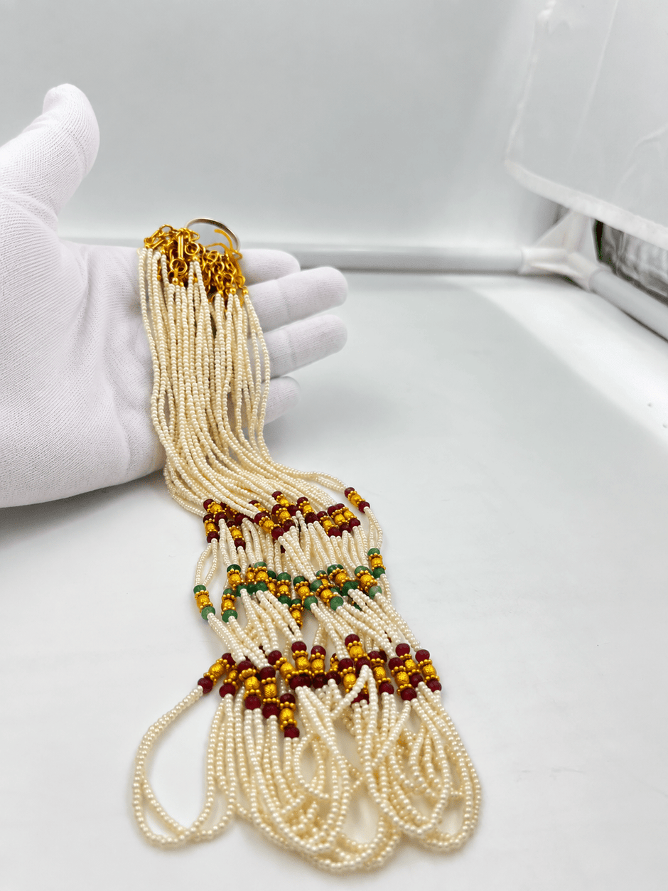 Double-strand white beaded necklace with deep red and green bead accents, decorative gold-tone elements and a gold-tone clasp displayed on a white necklace stand. - bhavmayi