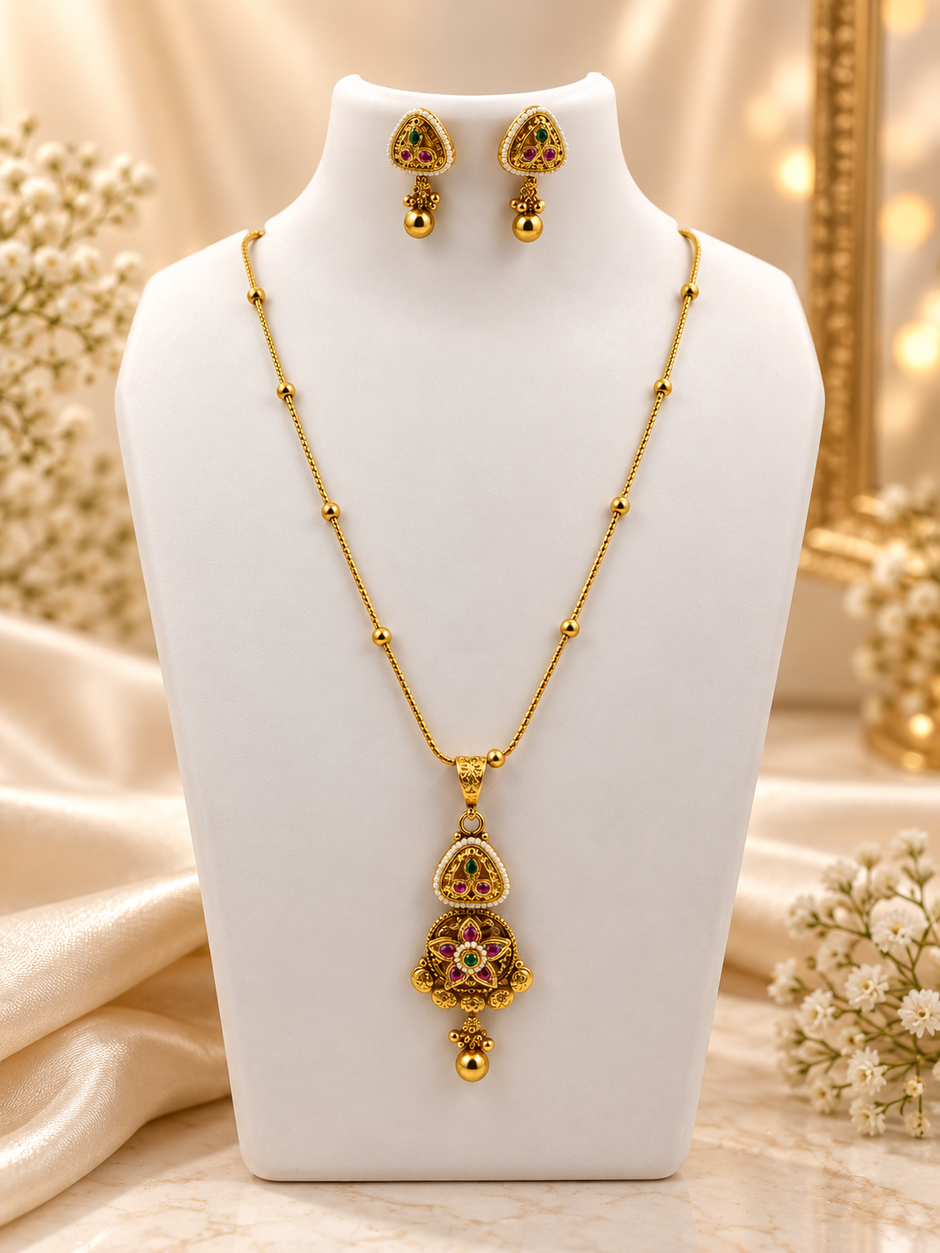 Gold-tone temple necklace set displayed on a white jewellery bust, featuring a decorative pendant with green and magenta accents, a beaded chain, and matching triangular drop earrings.
