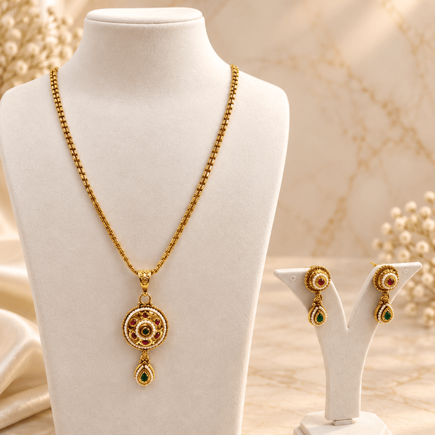 Gold-tone floral halo necklace set displayed on white jewellery stands, featuring a round pendant with green and magenta-pink accents, a decorative beaded chain, and matching earrings with green teardrop-shaped drops. - bhavmayi