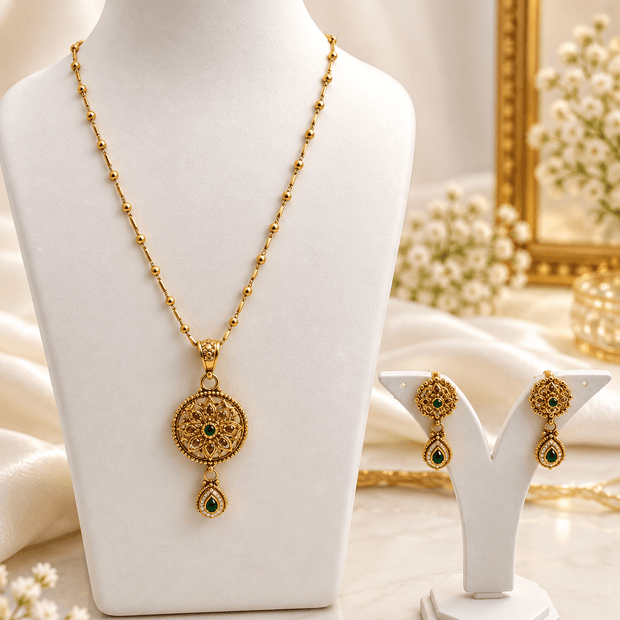 Gold-tone floral pendant necklace set displayed on white jewellery stands, featuring green and magenta-pink accents, a beaded chain, and matching earrings with green teardrop-shaped drops. - bhavmayi