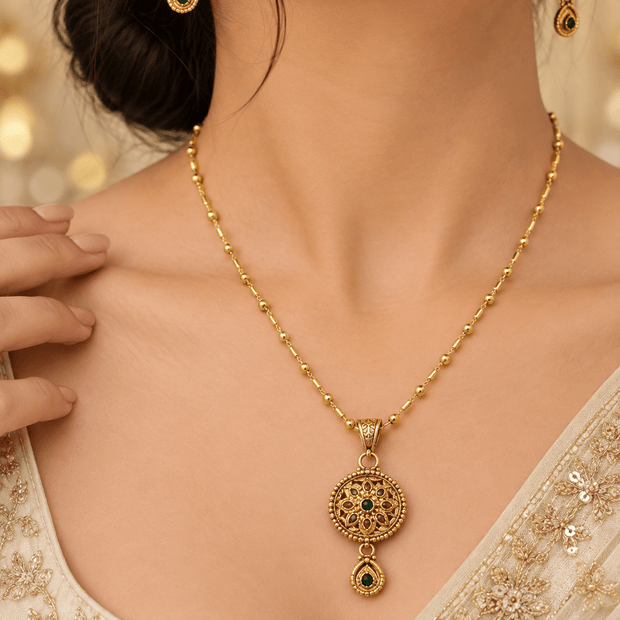 Gold-tone floral pendant necklace set displayed on white jewellery stands, featuring green and magenta-pink accents, a beaded chain, and matching earrings with green teardrop-shaped drops. - bhavmayi