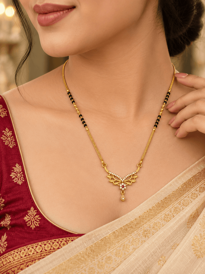Gold-tone mangalsutra necklace with black beads, symmetrical floral leaf-shaped centerpiece, clear decorative stones, red center accent, and dangling teardrop pendant on a white background. - bhavmayi