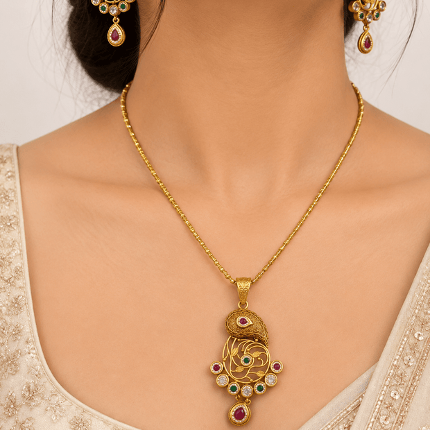 Gold-tone ornamental pendant and matching drop earrings displayed on a cream background, featuring intricate golden patterns, magenta-pink accents, green teardrop-shaped drops, and white decorative details. - bhavmayi