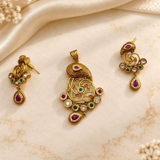 Gold-tone ornamental pendant and matching drop earrings displayed on a cream background, featuring intricate golden patterns, magenta-pink accents, green teardrop-shaped drops, and white decorative details. - bhavmayi