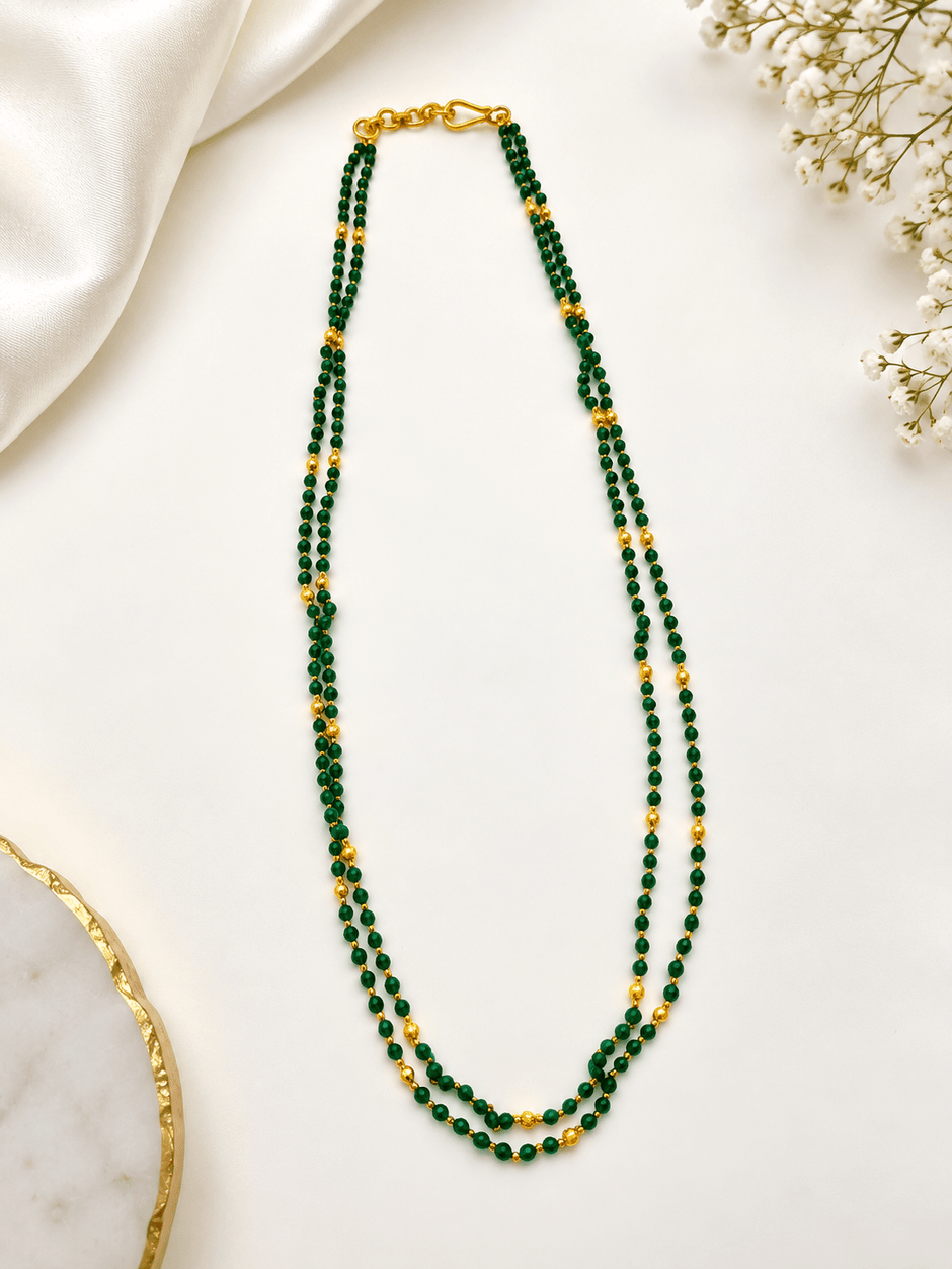 Green beaded necklace with small gold-tone connecting elements and a gold-tone clasp, displayed in a rounded elongated shape on a white jewellery display and against an elegant neutral background. - bhavmayi